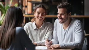 Two people are smiling and sitting at a table while listening to a third person. One person holds a pen and is ready to write, in a cozy indoor environment.