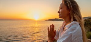 A person stands by the sea with folded hands and gazes into the distance. The sun is setting on the horizon, bathing the scene in a warm, golden light.