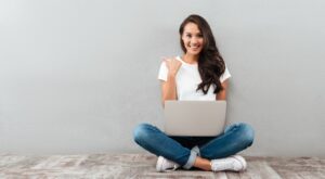 Eine Frau sitzt im Schneidersitz mit einem Laptop auf dem Scho&szlig; und l&auml;chelt, w&auml;hrend sie auf einen wei&szlig;en Hintergrund zeigt. Sie tr&auml;gt ein wei&szlig;es T-Shirt und blaue Jeans.