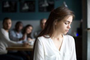 A woman with long brown hair sits pensively with her eyes closed, while four people sit around a table in the background and look at her. The scene is set in an interior room.