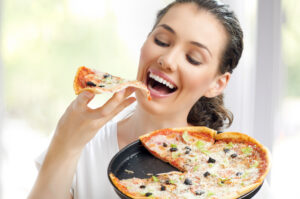 A woman laughs as she holds a slice of pizza and a pizza plate in her hands. She is about to bite into the slice. The background is blurred and bright.