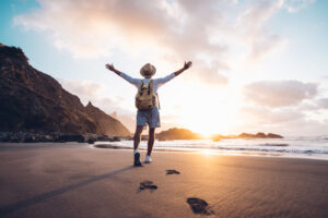 Eine Person mit Rucksack und Hut breitet die Arme aus, w&auml;hrend sie &uuml;ber einen sandigen Strand spaziert. Im Hintergrund sind felsige Klippen und ein sonniger Himmel bei Sonnenuntergang zu sehen.