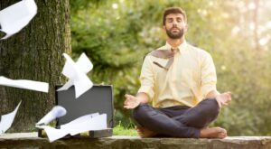 A man in a yellow shirt meditates cross-legged in front of a tree. Papers fly out of an open briefcase while he keeps his eyes closed, relaxed and surrounded by green nature.