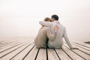 A couple sits tightly embraced on a wooden jetty, both in warm sweaters, as they gaze out over calm waters. The sky is bright and slightly cloudy.
