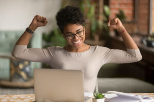 A person with glasses raises her arms triumphantly in front of a laptop. She smiles in a cozy room with brick walls and plants in the background. A small table with documents and a cactus is visible.