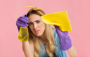A woman with long hair is wearing purple rubber gloves. She is holding a spray bottle with yellow liquid and a yellow cloth. The background is plain pink.
