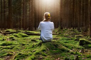Eine Person mit blonder Haarblume sitzt ruhig auf moosigem Boden in einem dichten Wald, umgeben von hohen B&auml;umen, w&auml;hrend sanftes Sonnenlicht hereinstr&ouml;mt und eine ruhige Atmosph&auml;re schafft.