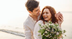 A woman and a man stand smiling on the beach, the woman holding a bouquet of white wildflowers. Both are relaxed and enjoying a sunny, peaceful moment by the water.