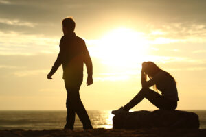 A man walks away while a woman sits on a rock and holds her head in her hands. Both people are depicted as silhouettes in the sunset on the beach.