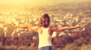 A person with outstretched arms stands with his back to the camera and looks out over a blurred cityscape at sunset, surrounded by soft, golden light and nature in the foreground.
