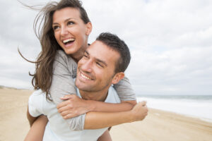 Eine Frau umarmt lachend einen Mann von hinten, der ebenso l&auml;chelt. Sie stehen an einem sandigen Strand. Der Himmel ist bew&ouml;lkt, und das Meer ist im Hintergrund sichtbar.