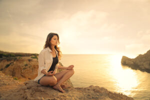 A person is meditating cross-legged on a rock. His gaze is directed thoughtfully towards the sea. The surroundings show a rocky coast at sunset with calm water.