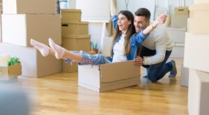 A woman sits enthusiastically in a cardboard box while a man cheerfully pushes her through a room full of moving boxes. Both show joy, surrounded by bright, modern furnishings.