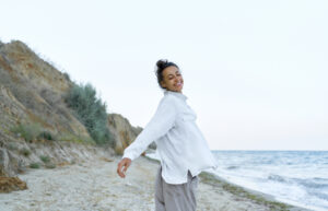 Woman in a white shirt stands smiling on a beach with her arms outstretched. The sea and a rocky cliff can be seen in the background. The sky is clear.