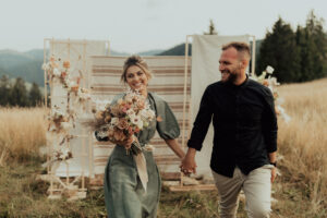 A smiling woman with a bouquet of flowers and a smiling man hold hands as they walk in front of decorative panels in a meadow-like setting with mountains in the background.