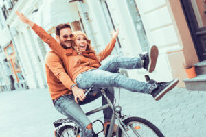 A man rides a bicycle with a woman sitting happily on the handlebars. Both are wearing casual clothes and laughing, surrounded by an urban street scene with bright buildings.
