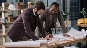 Two men in business attire study construction plans on a table in a modern office with shelves full of architectural models and safety helmets. Plants are visible in the background.