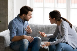 Two people are sitting on a gray sofa and gesticulating animatedly. Both seem to be engrossed in an intense conversation. In the background is a large window through which daylight is streaming in.