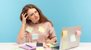 A person sits in front of a laptop covered in sticky notes, looking confused while scratching their head. In the foreground are colored paper, a pen and a smartphone. The background is plain light blue.