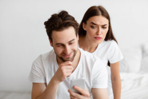Ein Mann h&auml;lt l&auml;chelnd ein Smartphone, w&auml;hrend eine Frau hinter ihm neugierig auf das Ger&auml;t blickt. Beide tragen wei&szlig;e T-Shirts und befinden sich in einem Schlafzimmer mit wei&szlig;en W&auml;nden.