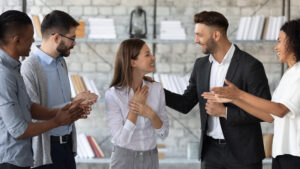 A group of five people are standing in a modern office. They clap and smile as they gaze appreciatively at a woman in the middle. Shelves of books line the brick wall.