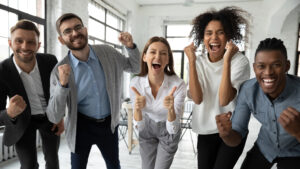 A group of five smiling people cheer and raise their fists. They are standing in a bright, modern room with large windows and white walls.