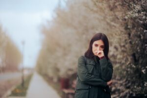 A woman with long dark hair, dressed in a green coat, stands thoughtfully on a sidewalk, surrounded by blossoming trees, while the background is blurred.