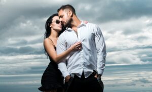 A couple stand in a tight embrace, the man in a white shirt and the woman in a black dress, against a dramatic, cloudy sky. Both are gazing dreamily into the distance.