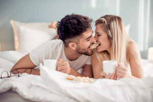 A couple lie smiling in bed, simultaneously biting into the same pastry and holding a cup. Soft bed linen and a bright, relaxed room surround them.