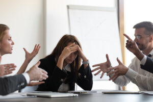Several people in formal dress are gesticulating and discussing at a table. A woman in the middle holds her head in her hands and looks stressed. There is a flipchart in the background.