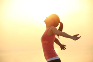 Woman with arms outstretched to the side stands in the sunset, head back. She is wearing a sleeveless top, while the light of the sky surrounds her.