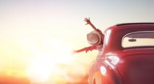 A red car drives off into the sunset. A person is leaning out of the window, joyfully stretching out their arms and wearing a straw hat. The sky is largely clear and bright.