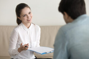 A woman in a white shirt holds a pen over a clipboard and smiles as she listens to a person across from her in a relaxed living room setting.