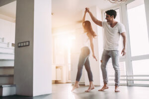 A couple dances happily barefoot in the sunlight of a modern living room. The woman spins while the man holds her hand. Stairs and large windows are visible.