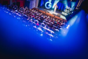 A person stands on a stage and speaks in front of a full audience in an auditorium. The scene is bathed in blue light. The text "Take you further" is visible.