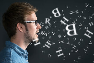 A man with glasses speaks while letters in various sizes and oblique arrangements stream out of his mouth against a dark background.