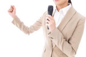 A person in a beige suit holds a microphone in his right hand, raises his left fist and speaks with commitment against a white background.