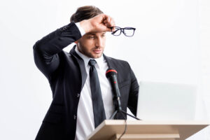 A man in a suit holds a pair of glasses in his right hand above his head in front of a microphone. He is standing at a desk, looking at a laptop screen, surrounded by bright light.