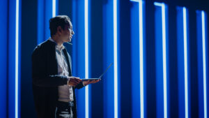A person holds a laptop and speaks on a stage. Vertical blue strips of light shine in the background, creating a modern, futuristic atmosphere.