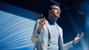 A person wearing glasses and a light-colored turtleneck sweater is giving a presentation, gesticulating with both hands and speaking into a microphone in front of a large-format screen with blue, curved lines in the background.