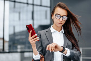 Eine Frau in Businesskleidung schaut auf ihre Armbanduhr, w&auml;hrend sie ein rotes Smartphone in der anderen Hand h&auml;lt. Im Hintergrund befindet sich ein modernes Glasgeb&auml;ude.
