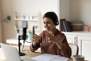 A woman with headphones speaks into a microphone in front of a laptop. She gestures with her hands. The room is a cozy home office with a wall shelf, books and a cup.