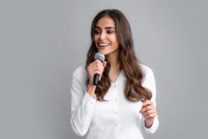 A smiling woman holds a microphone while standing in a gray, neutral background. She is wearing a white shirt and appears to be giving a speech.