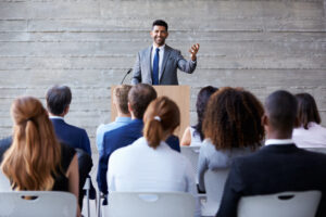 A man in a suit speaks with a smile at a wooden desk, surrounded by a microphone in a room with a concrete wall. Several people sit in rows in front of him and listen.