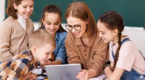 Person mit Brille und vier Kinder betrachten gemeinsam ein Tablet. Die Gruppe scheint fr&ouml;hlich und konzentriert. Im Hintergrund eine gr&uuml;ne Tafel, die auf ein Klassenzimmer hinweist.