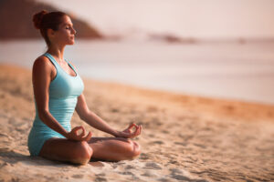 Eine Person sitzt in meditativer Haltung mit geschlossenen Augen am Strand. Sie tr&auml;gt ein hellblaues Sportoutfit, w&auml;hrend die sanften Wellen des Meeres im Hintergrund zu sehen sind.