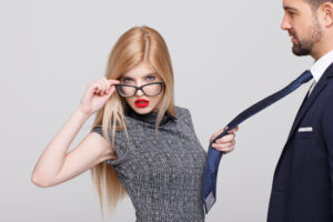 Eine Frau mit blonden Haaren und roten Lippen h&auml;lt eine Brille und den Schlips eines Mannes. Der Mann tr&auml;gt einen Anzug. Der Hintergrund ist einfarbig grau.