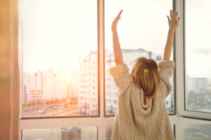 Eine Person mit langen Haaren streckt sich vor einem gro&szlig;en, offenen Fenster. Sonnenlicht str&ouml;mt herein und beleuchtet eine Stadtansicht mit modernen Wohngeb&auml;uden im Hintergrund.