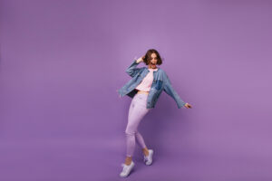 Eine Person tr&auml;gt eine blaue Jeansjacke und lila Jeans, steht in einer dynamischen Tanzpose. Der Hintergrund ist einfarbig violett, was den energetischen Ausdruck betont.