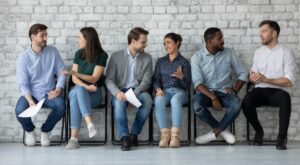 Sechs Personen sitzen auf St&uuml;hlen vor einer wei&szlig;en Backsteinwand und unterhalten sich miteinander. Einige halten Papier in den H&auml;nden, tragen Freizeitkleidung und wirken interessiert. Der Boden ist aus Holz.